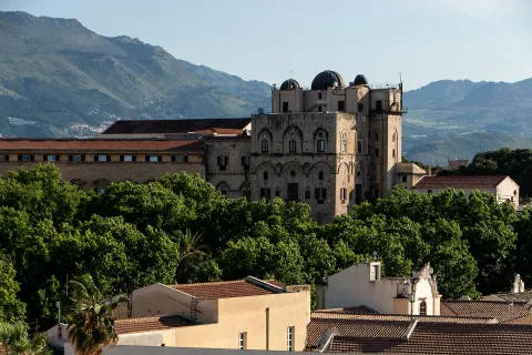 Panoramic view of the Palazzo dei Normanni in Palermo surrounded by trees with mountains in the background.